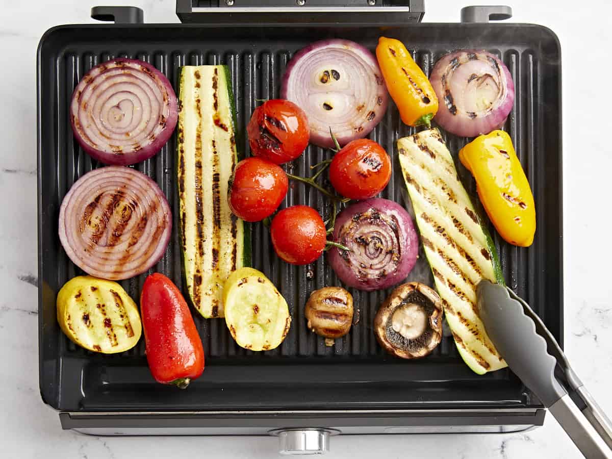 Grilled vegetables on a grill being flipped with tongs.