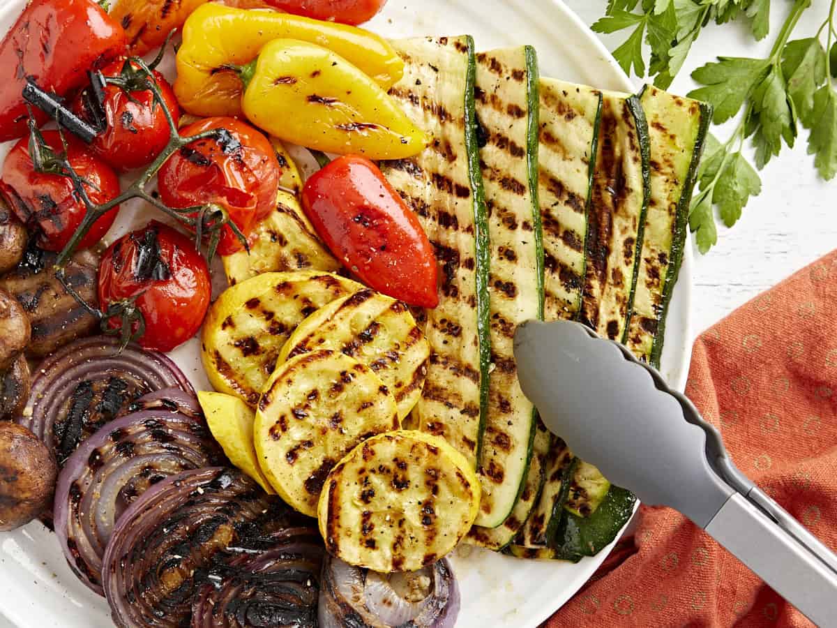 Overhead view of a plate of grilled vegetables with some tongs grabbing the zucchini.
