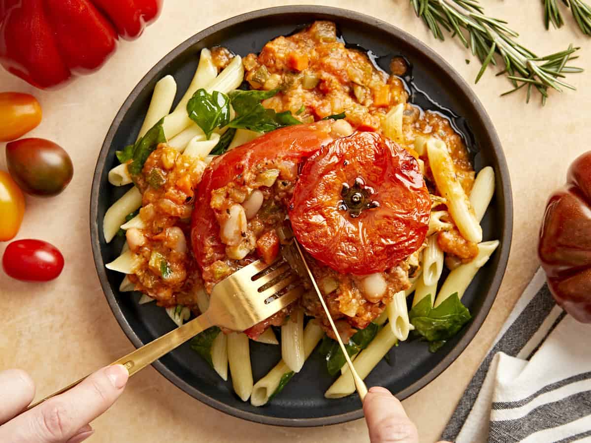 A knife and fork cutting into a tuscan white bean stuffed tomato on a plate with pasta.