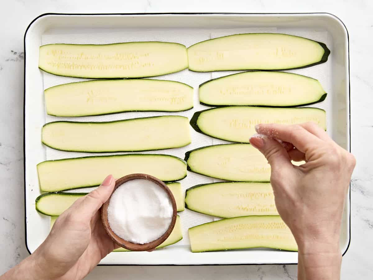 Slices of zucchini being sprinkled with salt in a baking dish