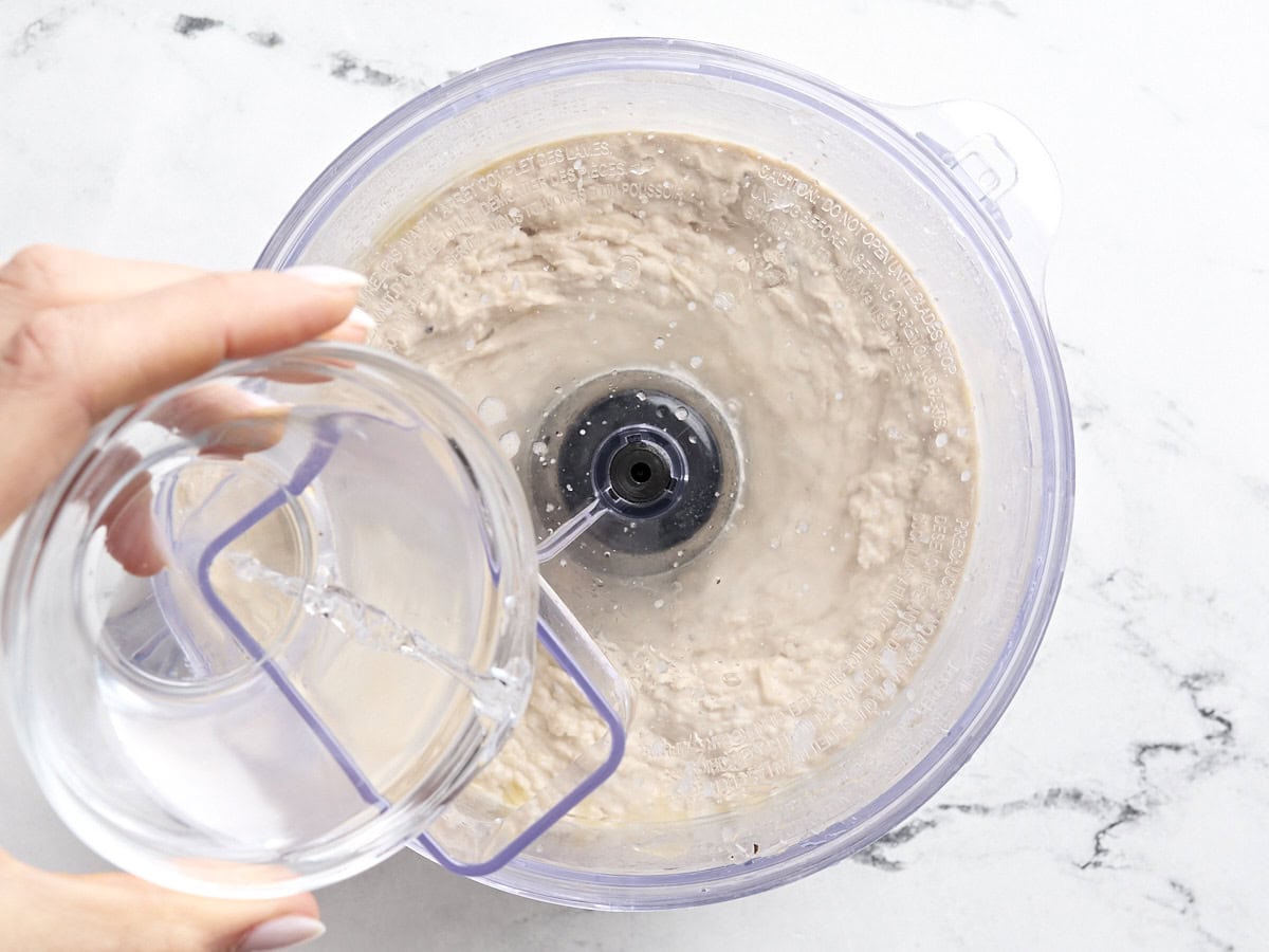 A hand pouring water into a food processor making white bean hummus