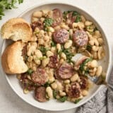 Overhead view of chicken sausage and white beans on a serving dish with toasted bread on the side and a fork lifting some out.