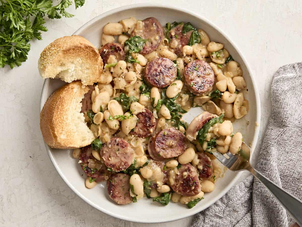 Overhead view of chicken sausage and white beans on a serving dish with toasted bread on the side and a fork lifting some out.