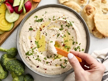 Close up of a hand dipping a carrot stick into a bowl of white bean hummus