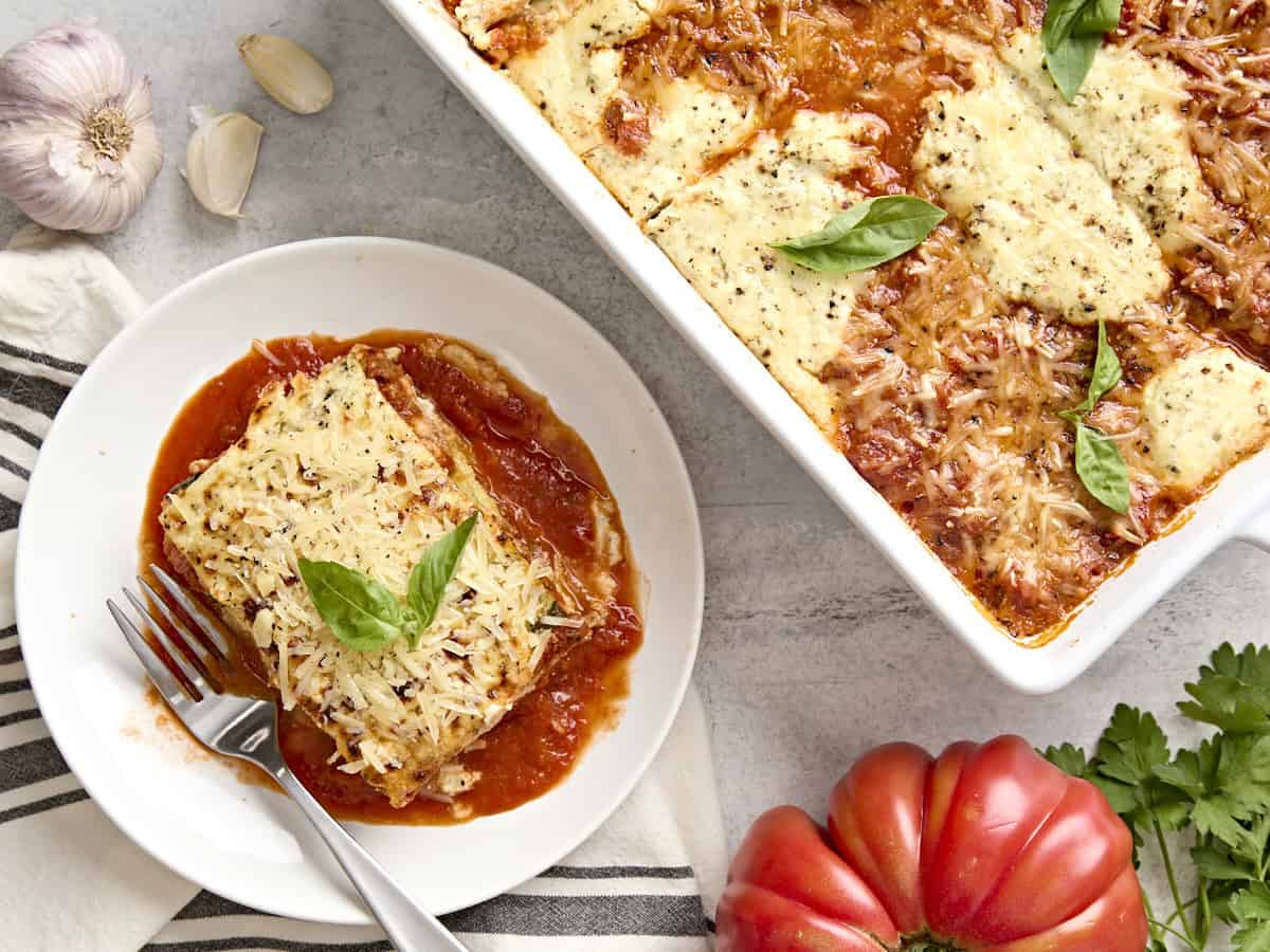 A slice of zucchini lasagna on a plate next to a baking dish filled with more zucchini lasagna