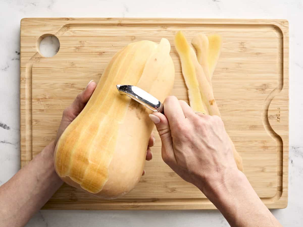 A hand peeling a butternut squash with a vegetable peeler.