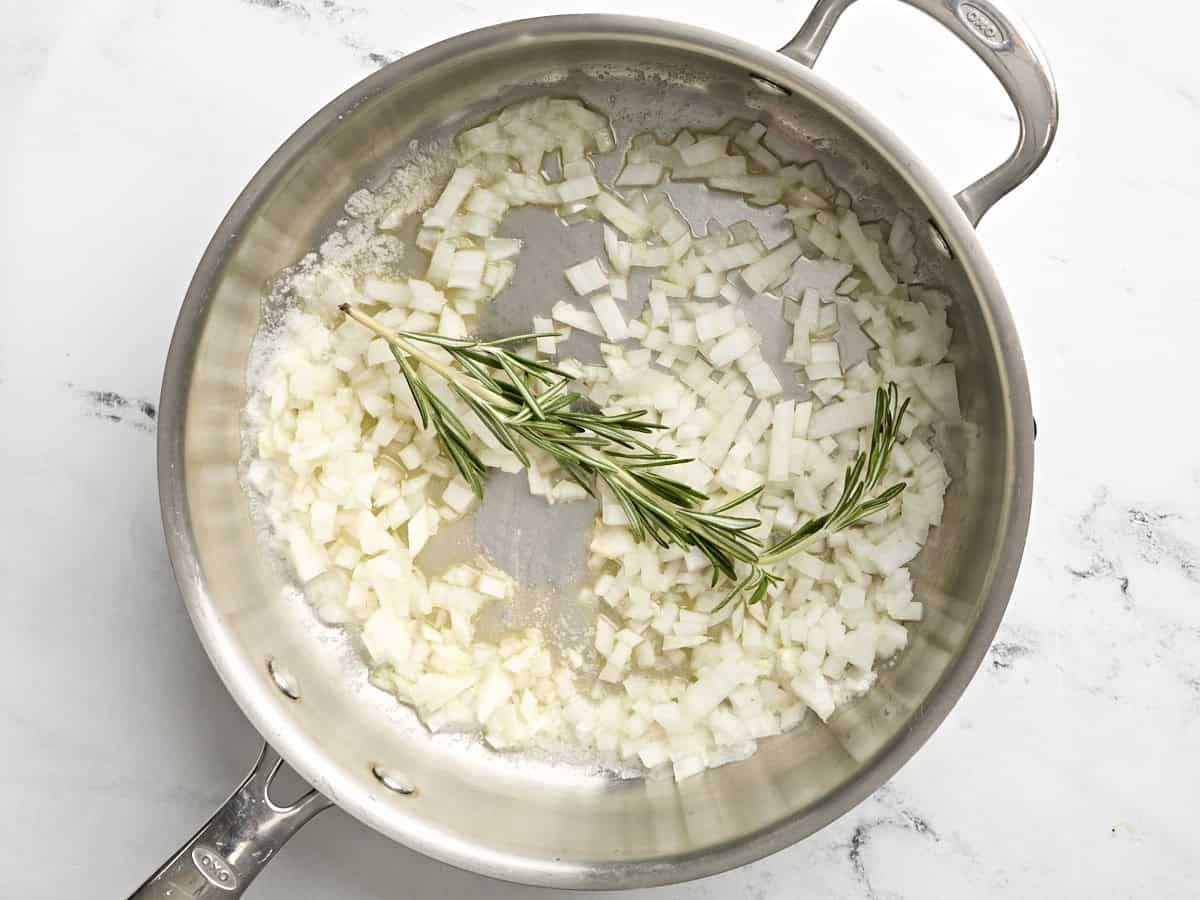 Diced onion and a sprig of rosemary sauteing in a skillet.