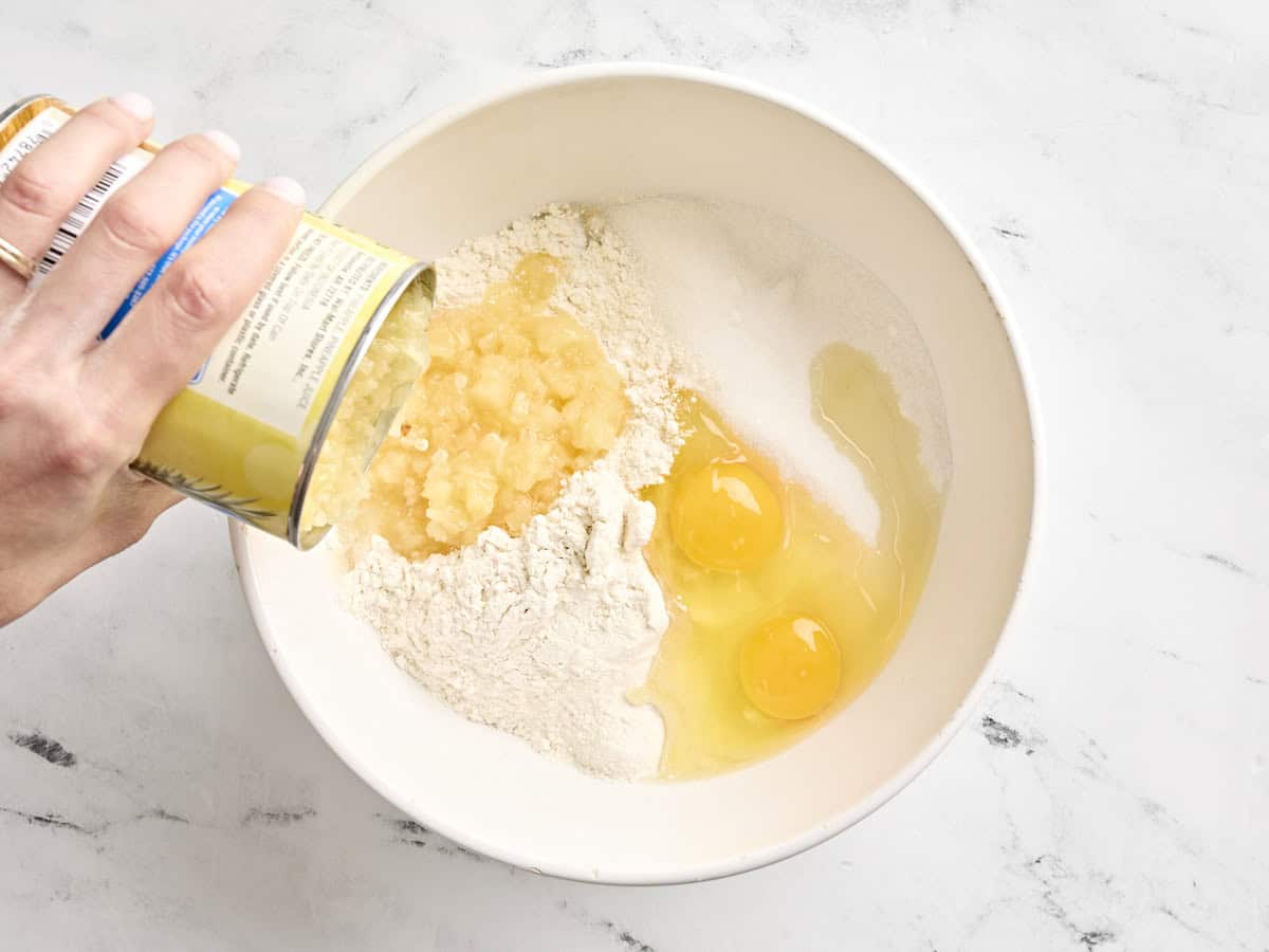 A can of crushed pineapple being added to a mixing bowl with eggs and flour.