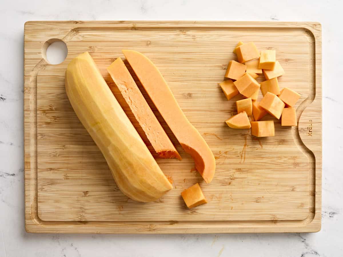 A butternut squash sliced in half, with one half sliced into cubes on a wooden chopping board.