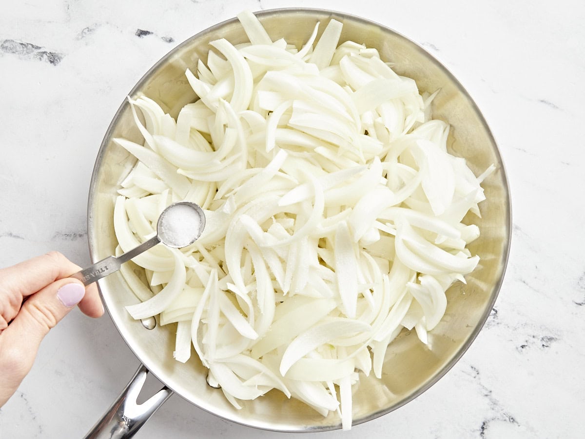 A hand holding a spoon of salt above sliced onions in a pan