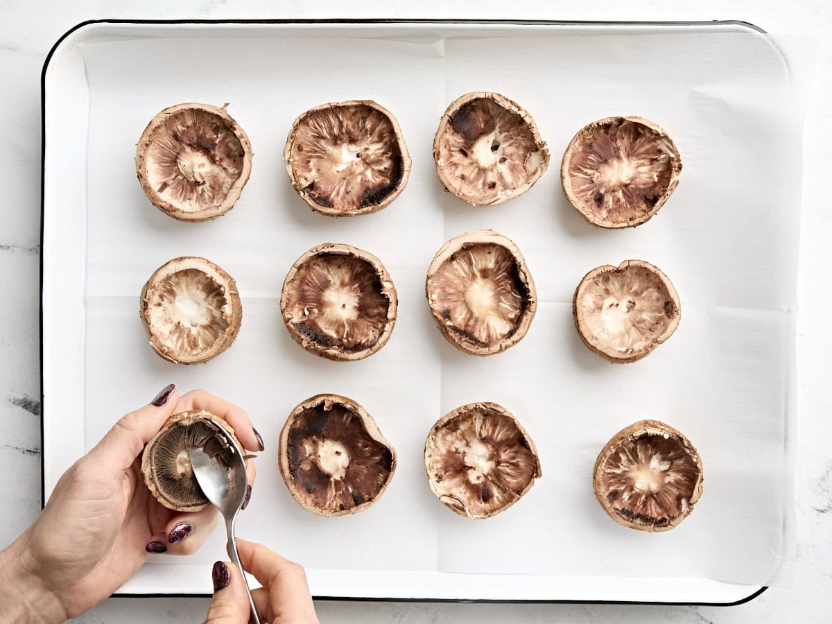 Mushroom caps on a baking sheet with a spoon scooping out the insides for stuffing.