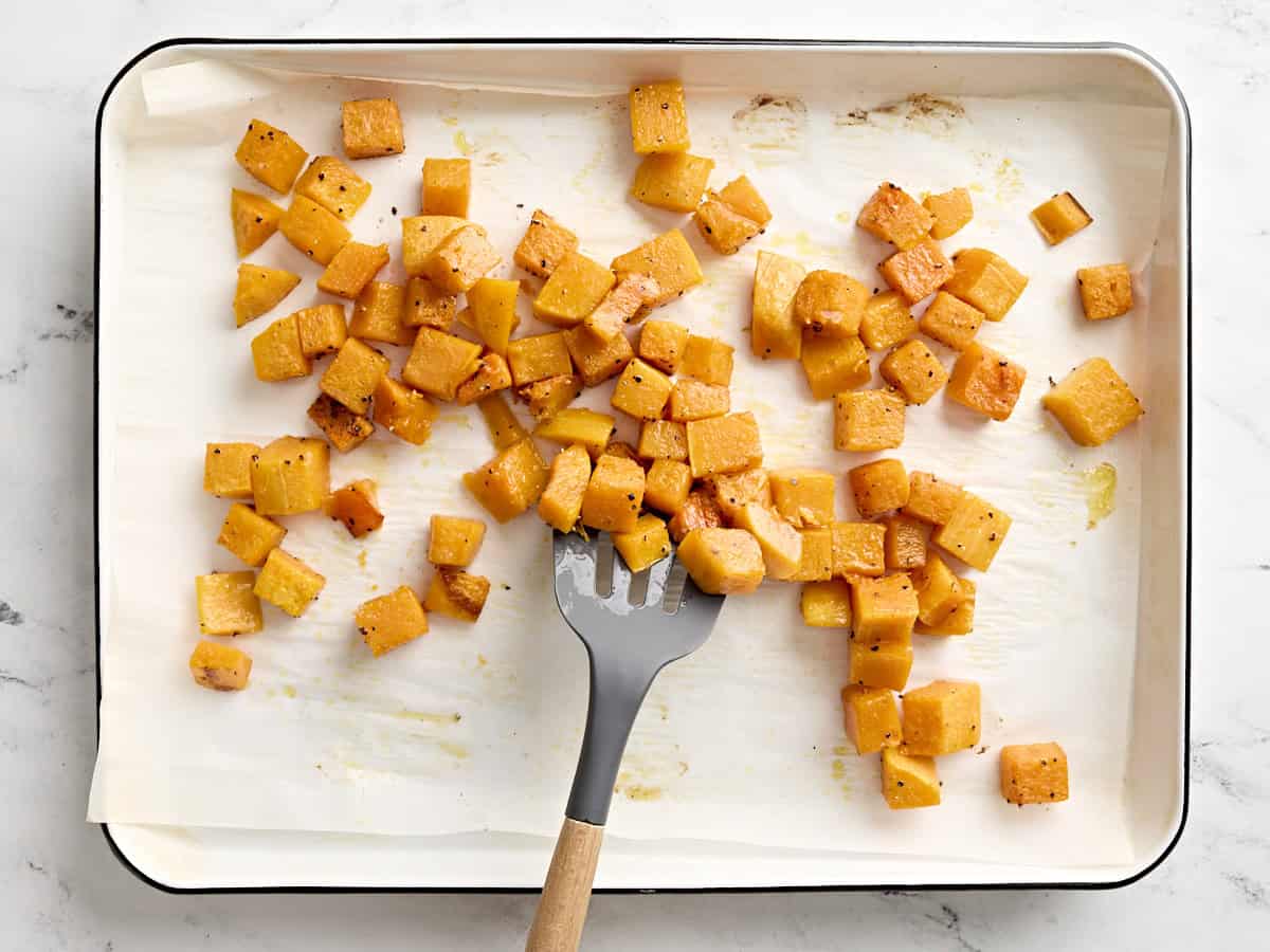 A spatula flipping roasted butternut squash over during cooking on a parchment lined baking sheet.