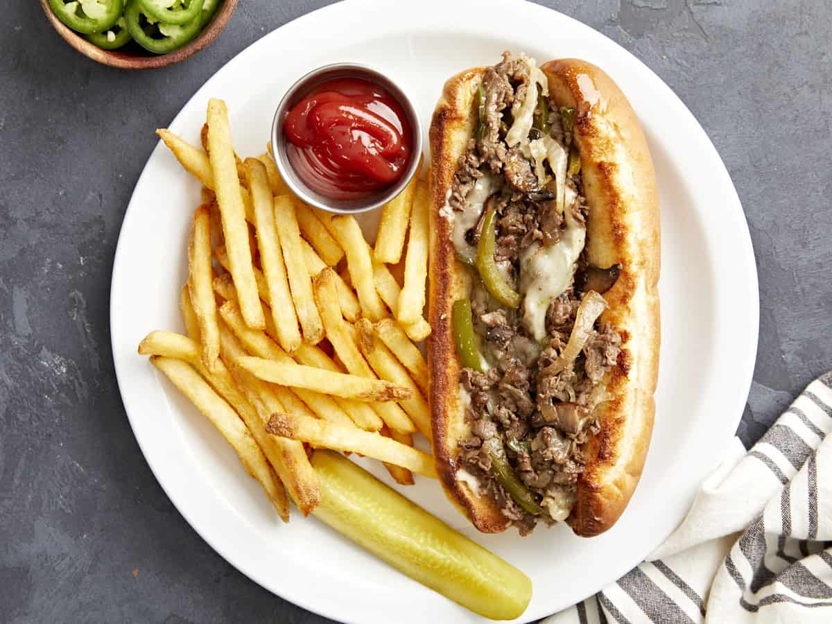 One Philly cheesesteak on a white plate with fries, ketchup, and a pickle on the side.