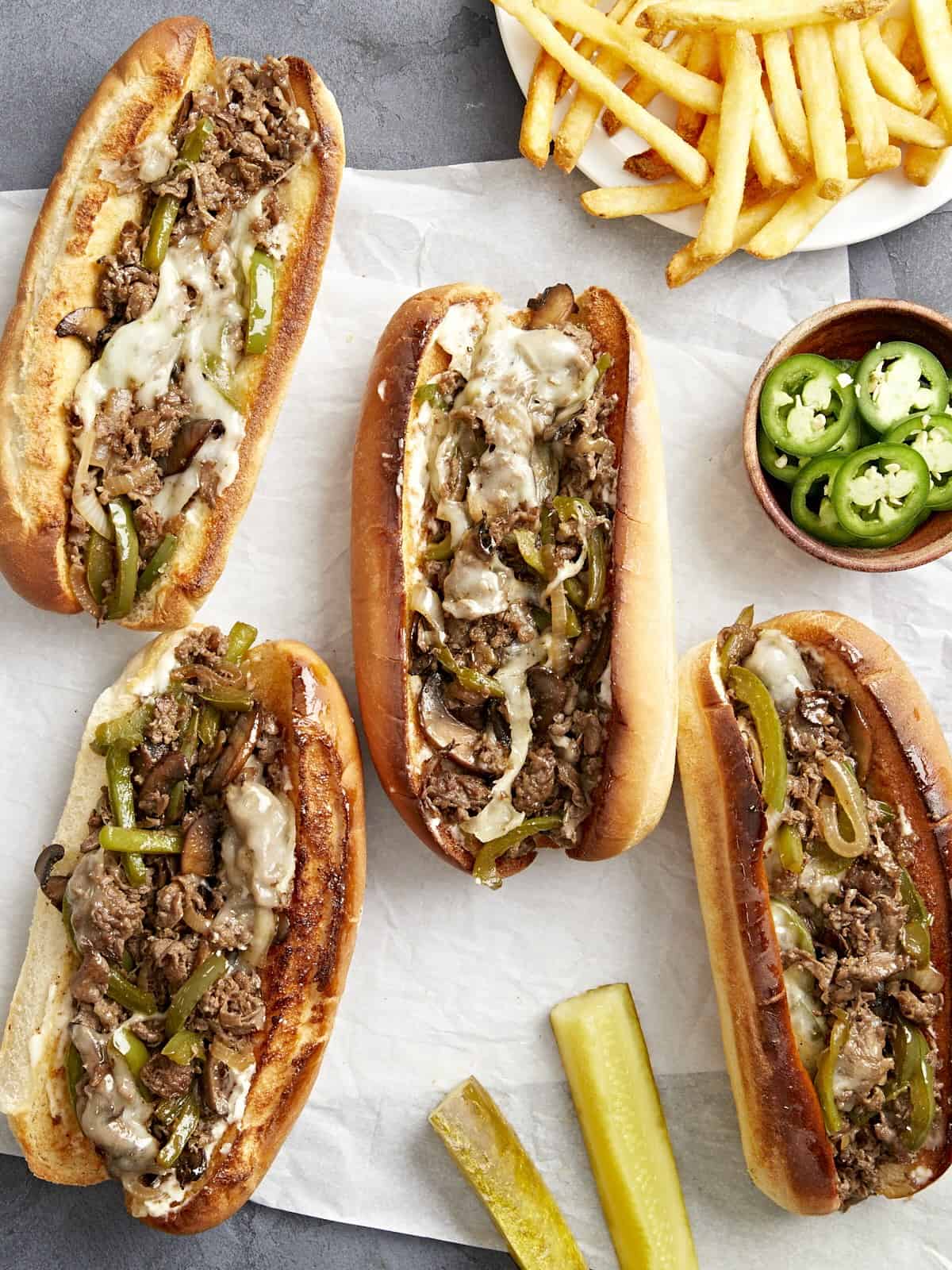 Overhead view of four philly cheesesteaks on parchment paper with fries and pickles on the side.