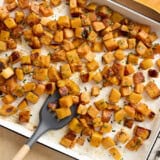 Overhead view of roasted butternut squash cubes on a baking tray with a spatula.