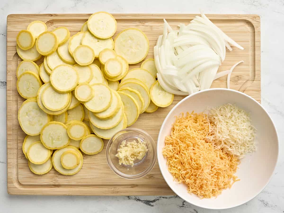 Sliced squash, onions, minced garlic, and shredded cheeses on a wooden chopping board.