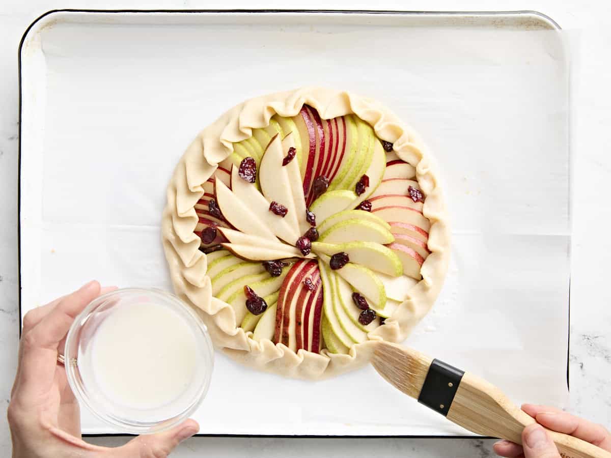 An unbaked pear galette being brushed with milk.