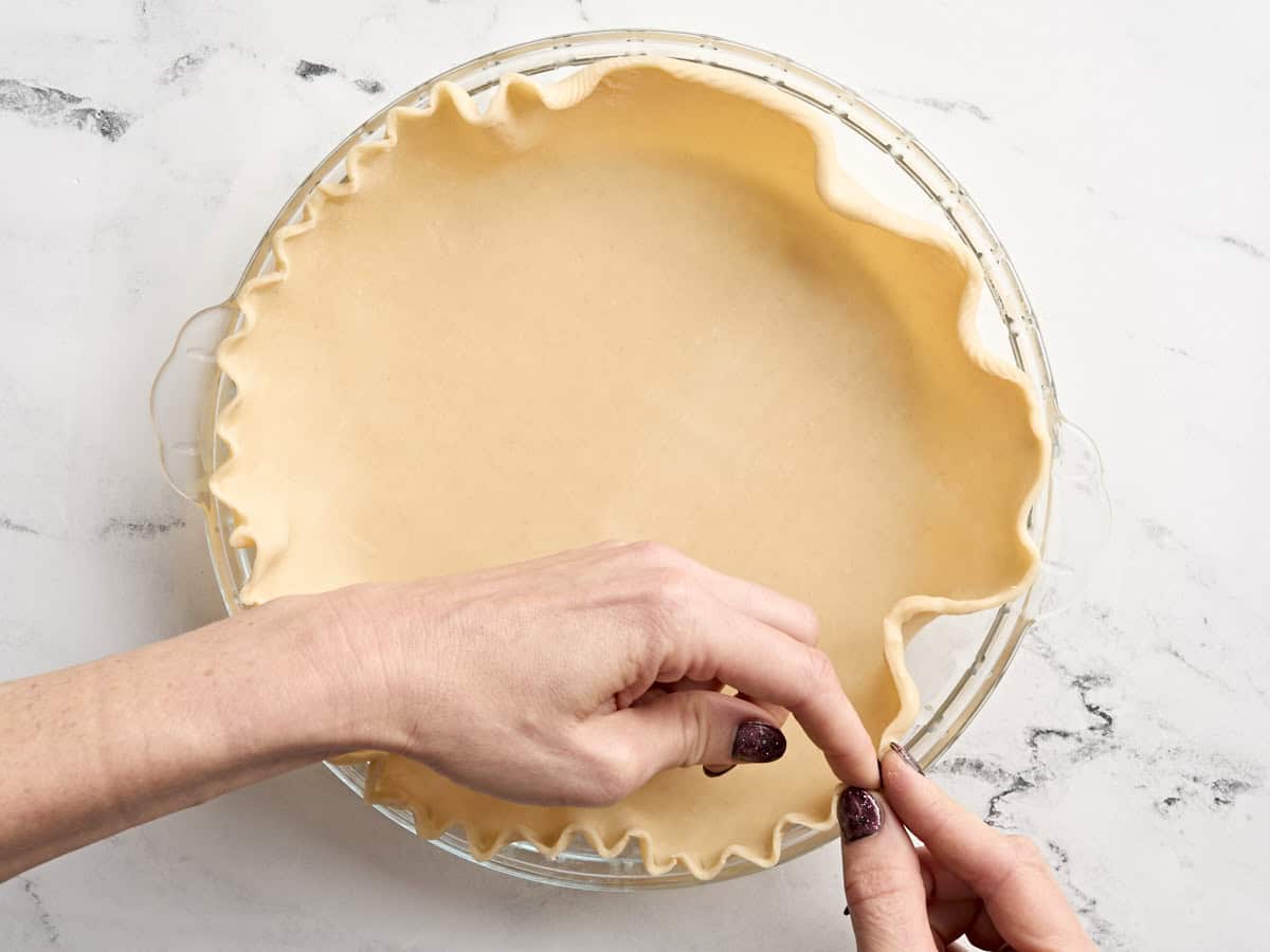 A hand crimping the edges of a pie crust in a pie dish for pecan pie.