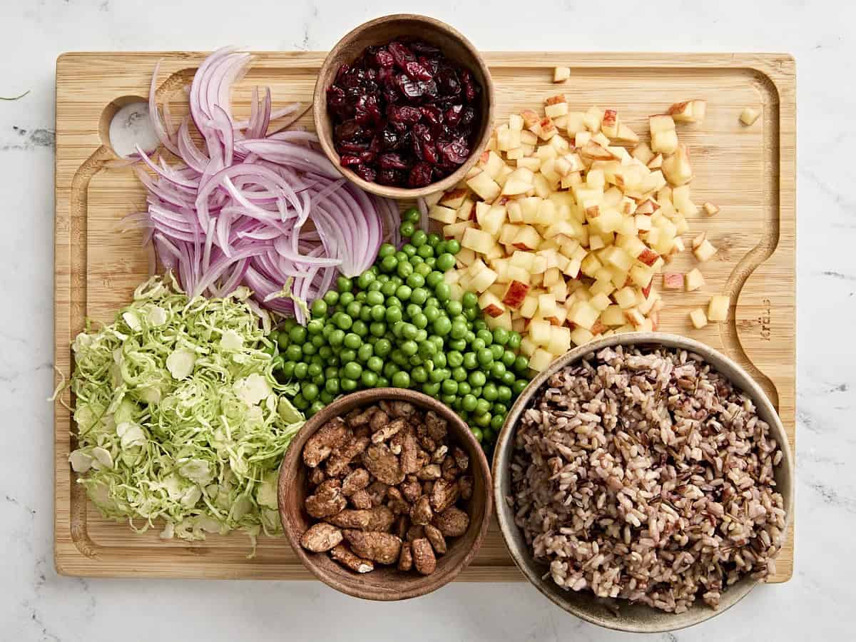 Sliced onions, shaved sprouts, chopped apple, dried cranberries, cooked wild rice, candied pecans, and frozen peas on a chopping board.