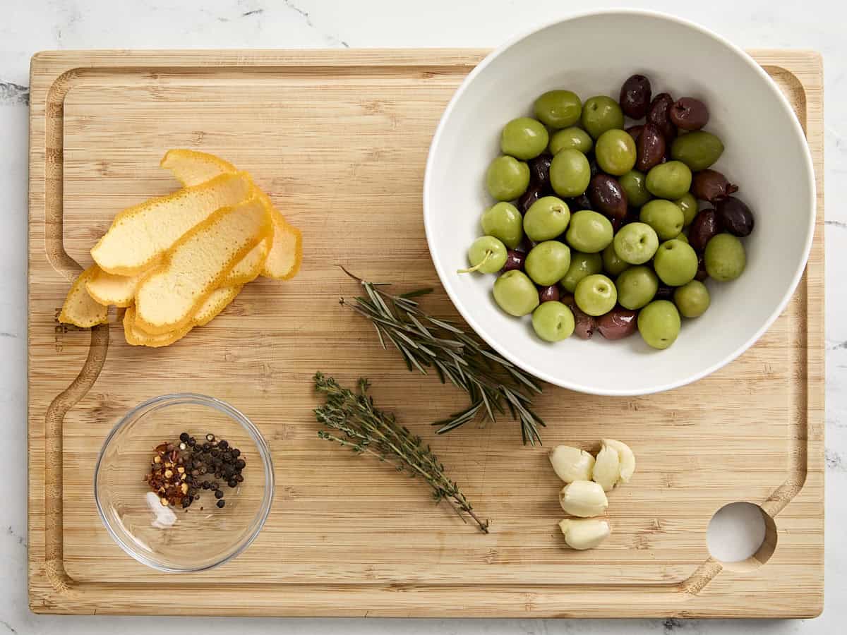 Overhead view of a bowl of olives, orange peel, fresh herbs, garlic cloves, and spices on a wooden chopping board.