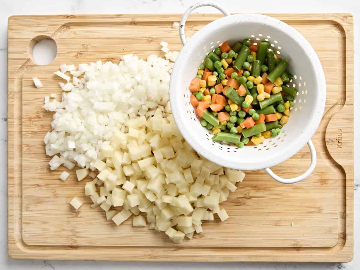 Diced onion, diced potato, and frozen vegetables on a wooden chopping board.