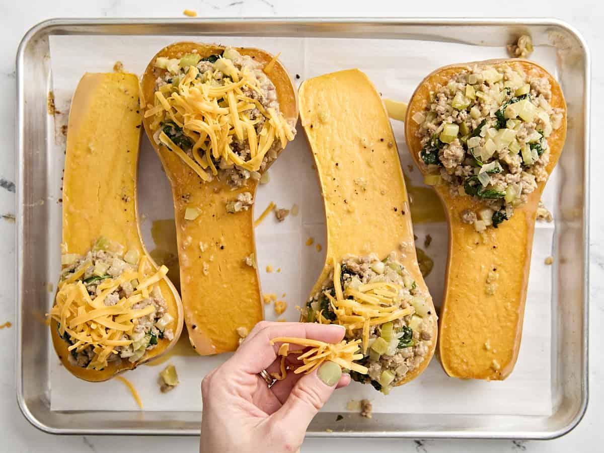 A hand sprinkling stuffed butternut squash with shredded cheese.