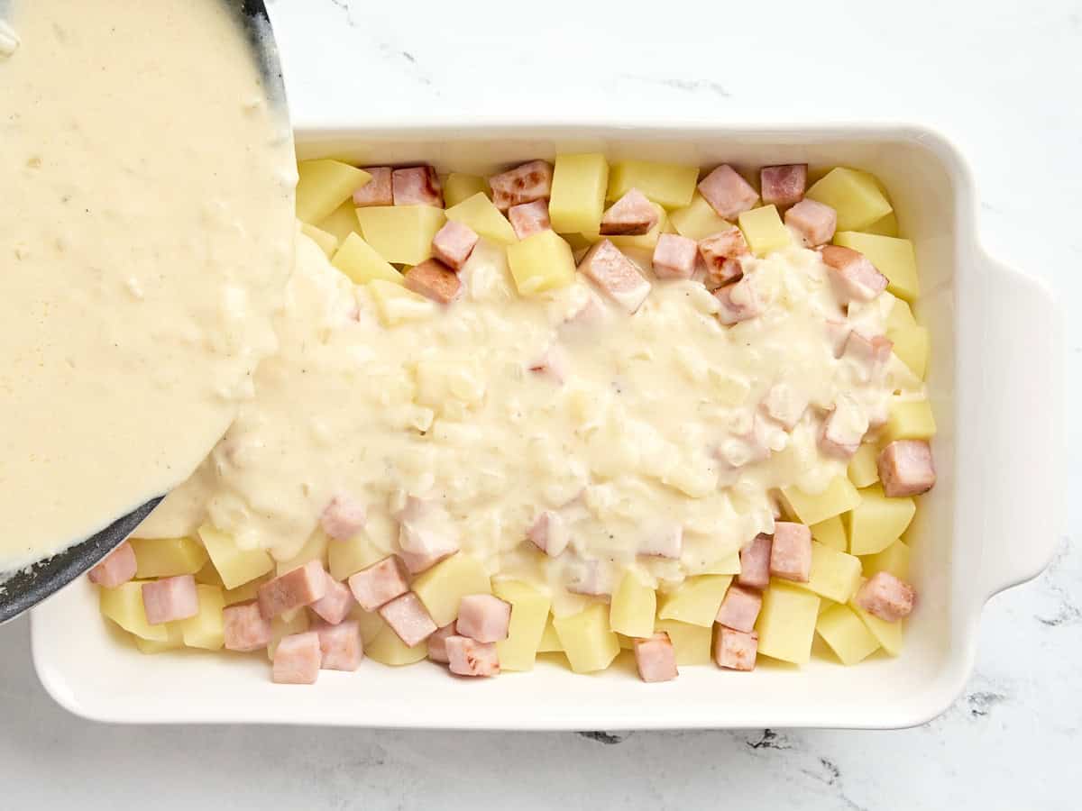 Cheese sauce being poured over diced potatoes and ham steak in a casserole dish.