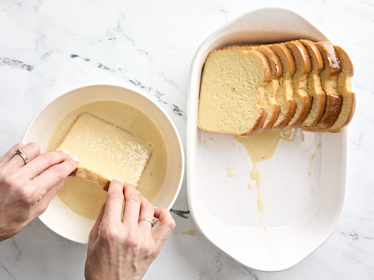 A hand dipping a slice of brioche into a bowl of egg mixture, next to a casserole dish with slices of dipped bread layered in.