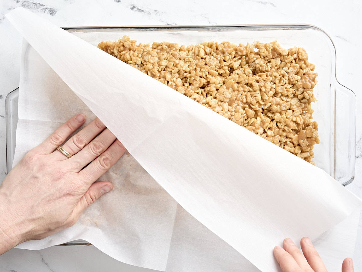 Hands pressing down the rice krispie mixture for Scotcharoos into a glass baking dish using a piece of parchment.