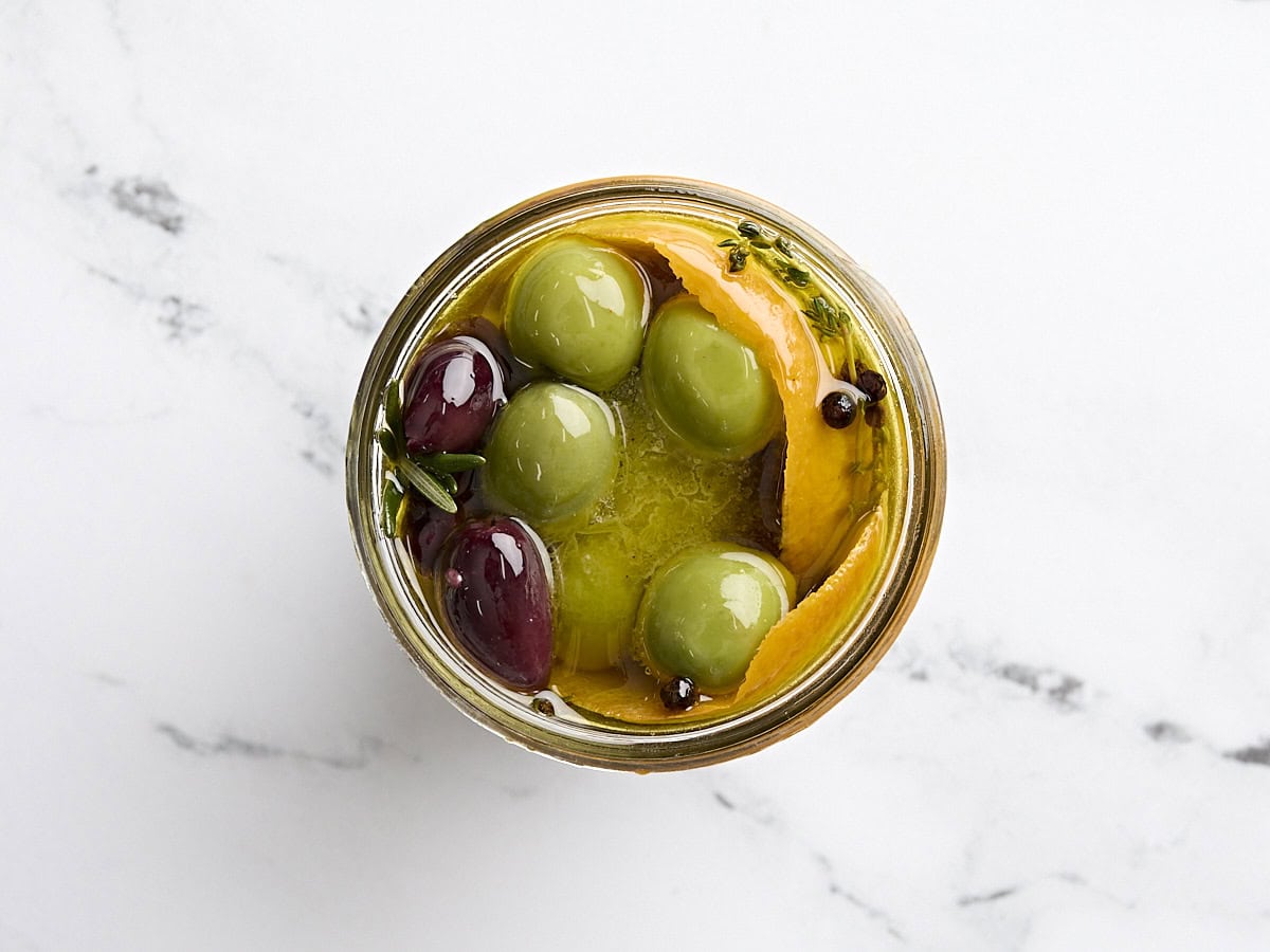 Overhead view of marinated olives with fresh herbs and orange peel in a mason jar.