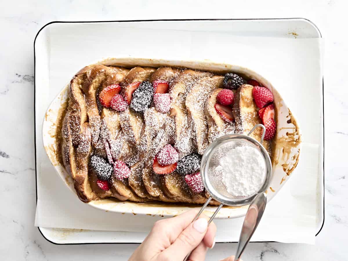 A hand sprinkling powdered sugar over baked fresh toast and berries in a casserole dish.