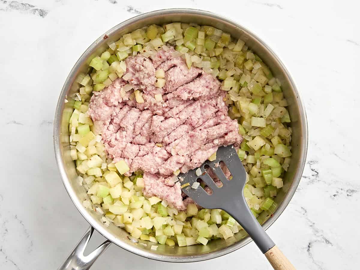 Ground sausage added to softened vegetables in a pan, being broken up by a spatula.