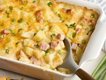 A serving spoon scooping ham and potato casserole from a baking dish.