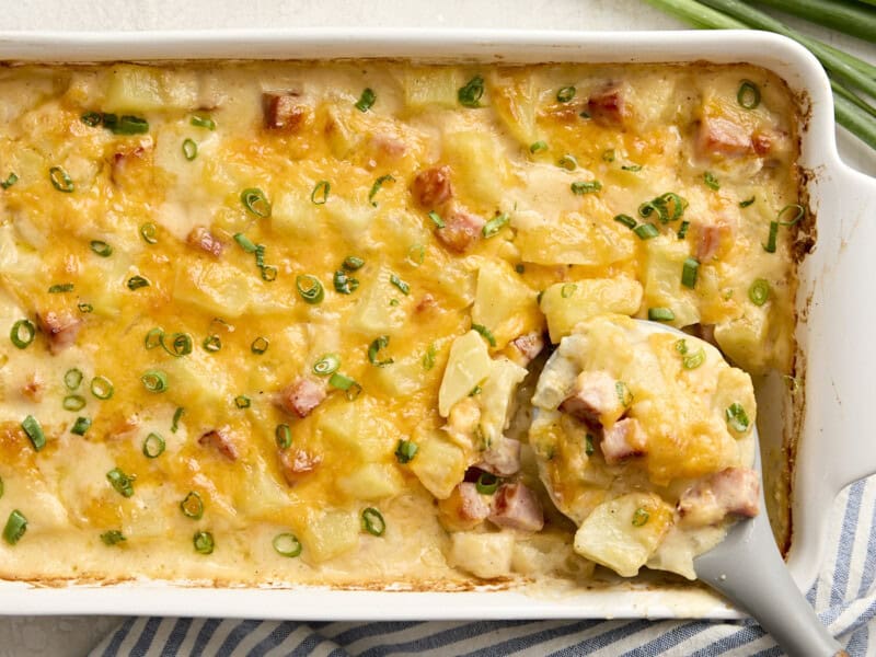 Overhead view of a serving spoon scooping ham and potato casserole from a baking dish.