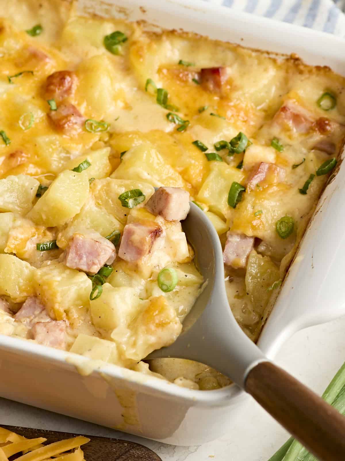 A serving spoon scooping ham and potato casserole from a baking dish.