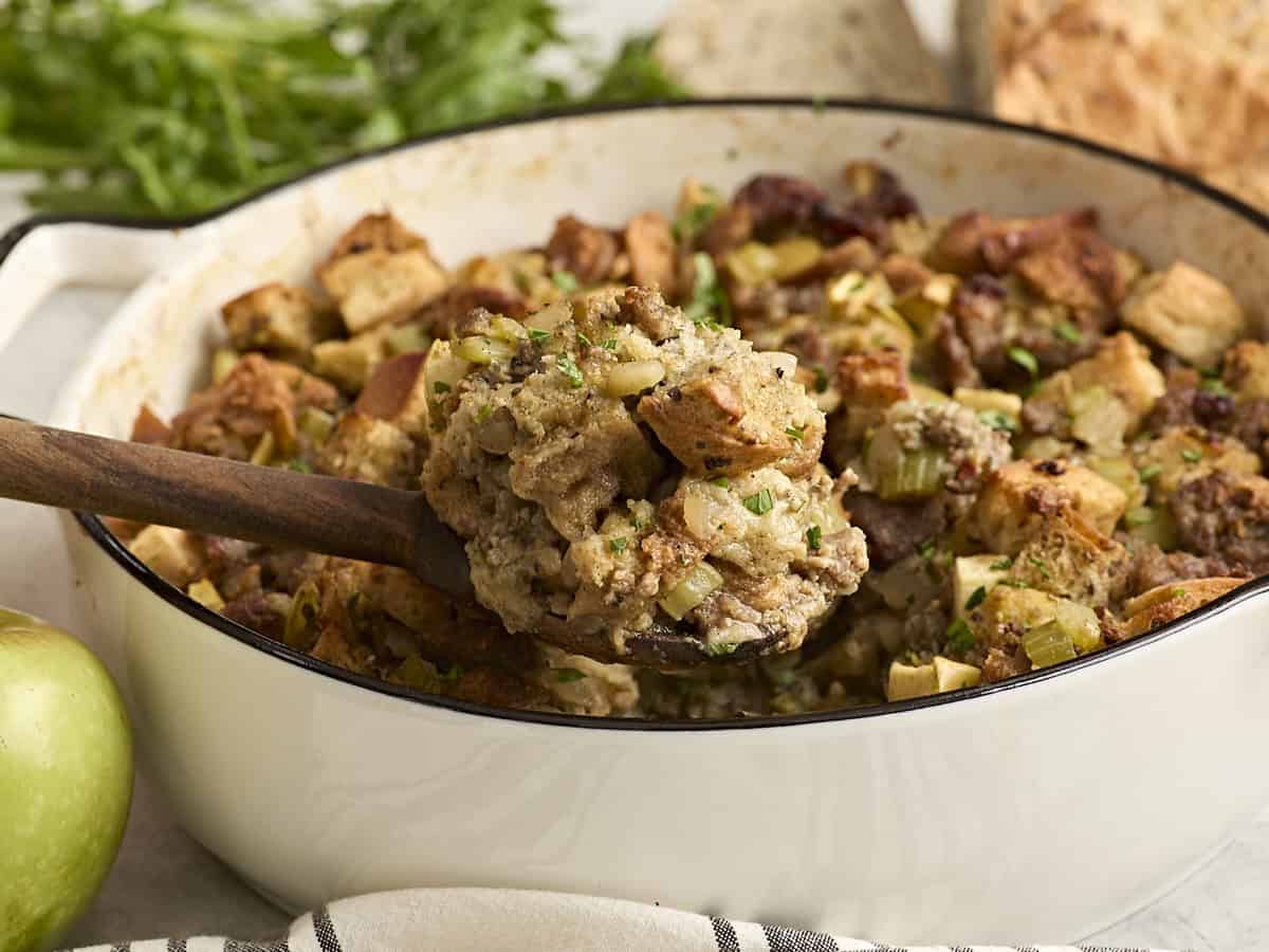 A wooden spoon scooping sausage stuffing from a casserole dish.