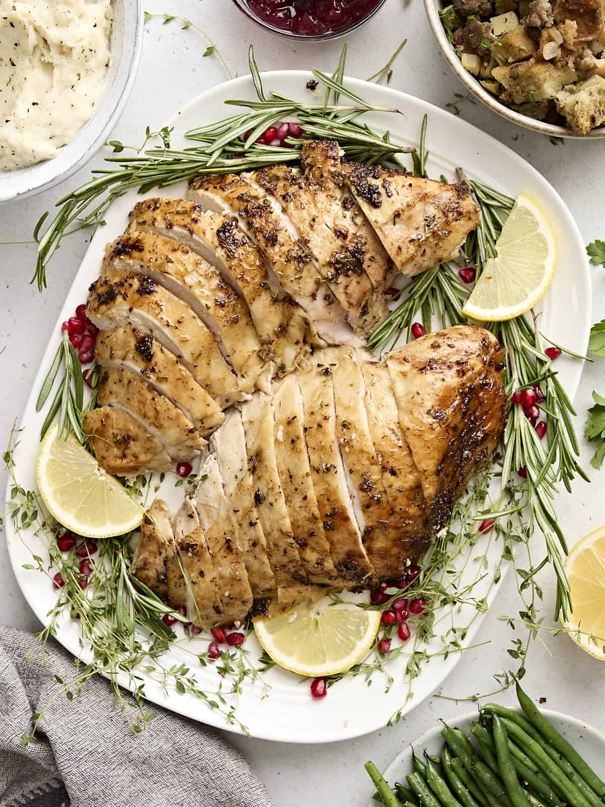 Overhead view of sliced turkey breast on a platter with fresh herbs and lemon slices as a garnish.