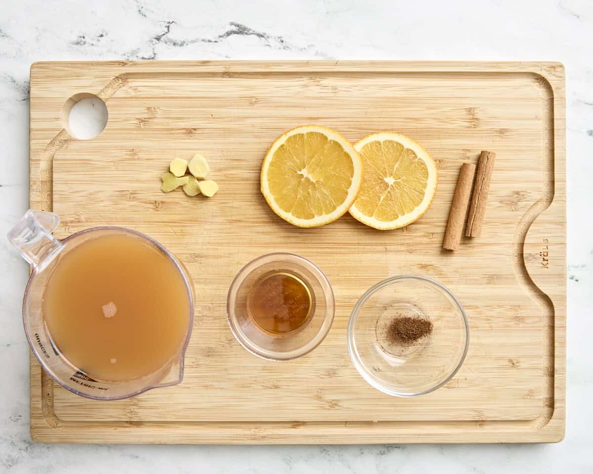 Apple cider, orange slices, chopped fresh ginger, maple syrup, nutmeg, and cinnamon sticks on a wooden chopping board.