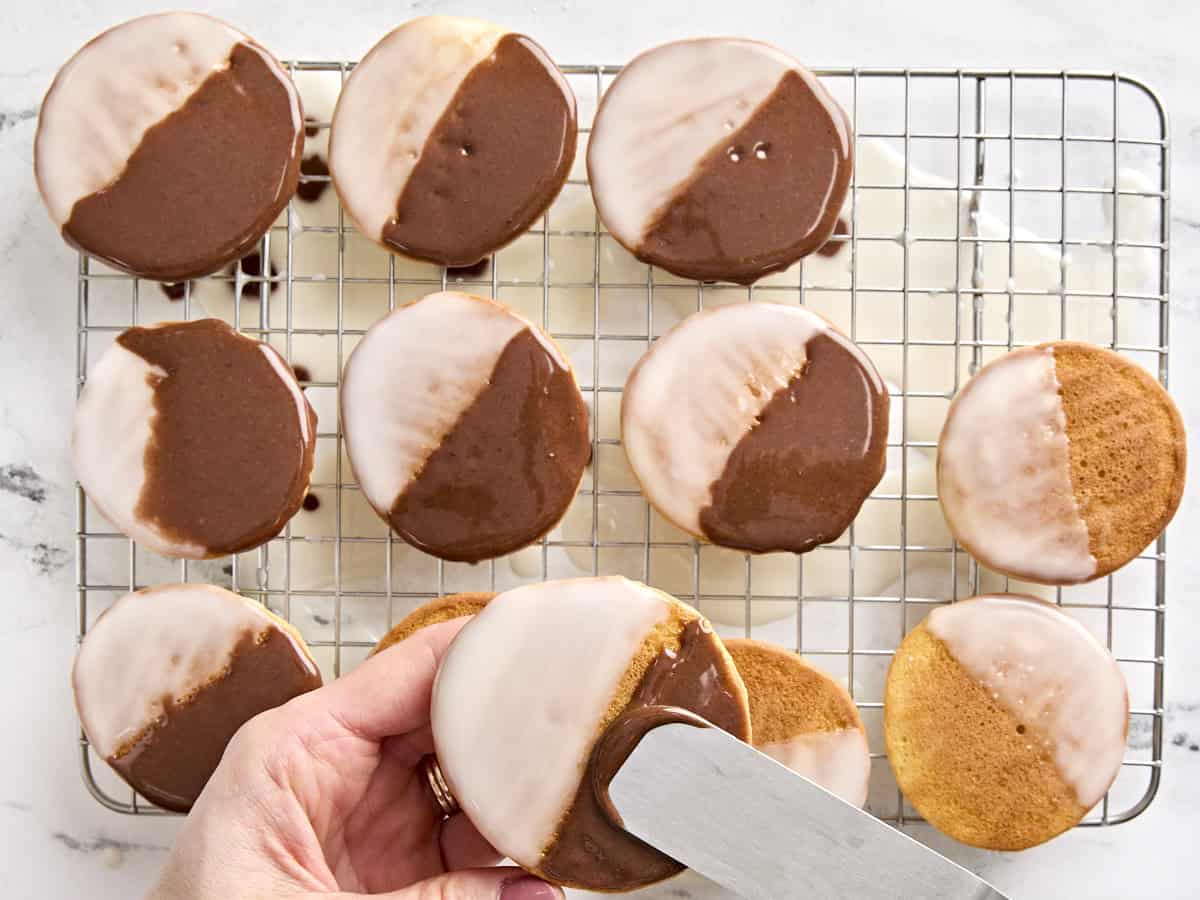 Black icing being added to black and white cookies with a butter knife.