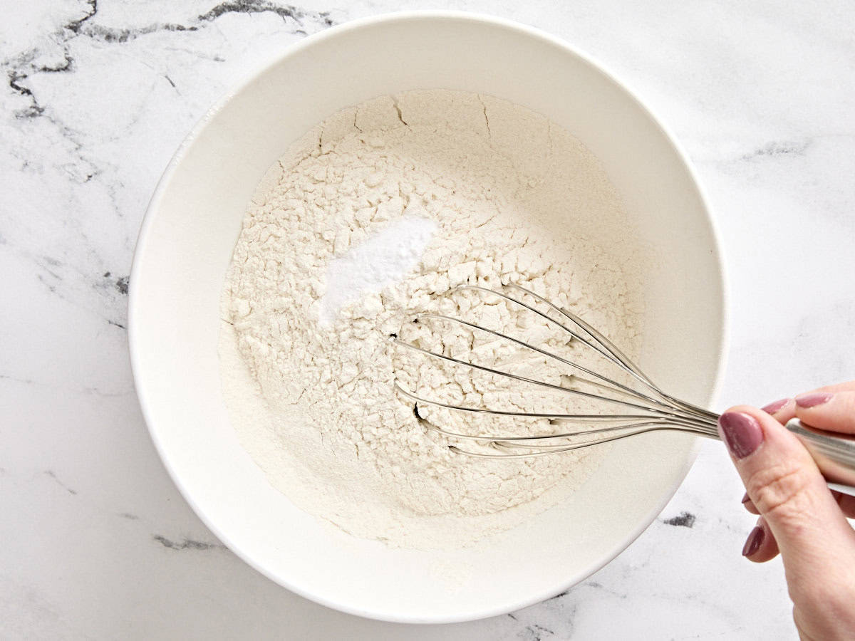 Baking soda and salt being whisked into sifted flour in a bowl.