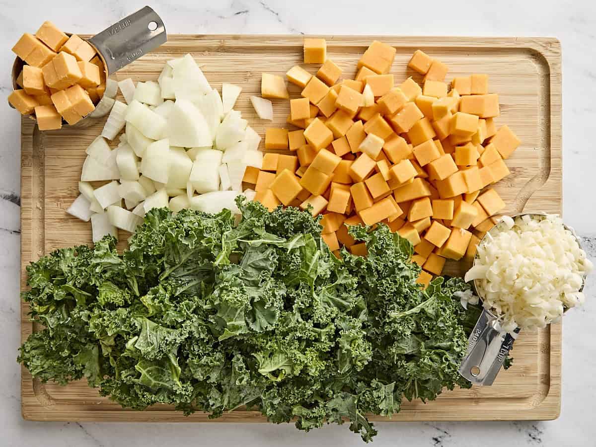 Chopped kale, diced butternut squash, cubed cheese, diced onion, and shredded mozzarella on a wooden chopping board.