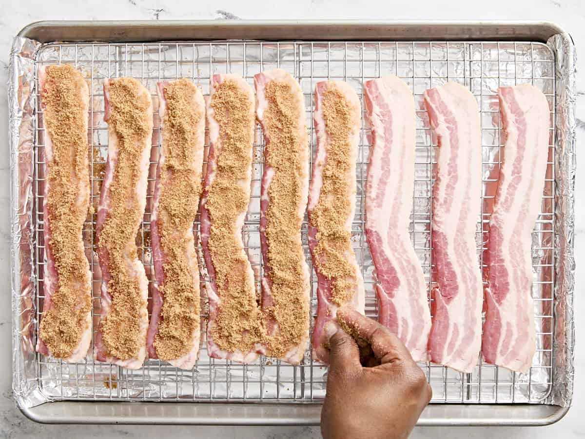 A hand sprinkling brown sugar over slices of bacon to make candied bacon.