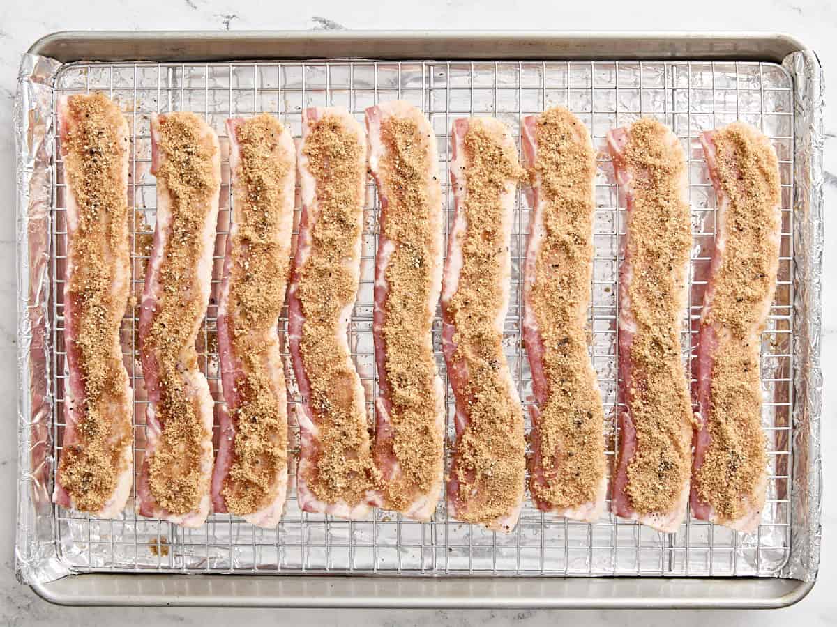 Slices of bacon on a wire rack over a baking sheet, topped with brown sugar and black pepper to make candied bacon.