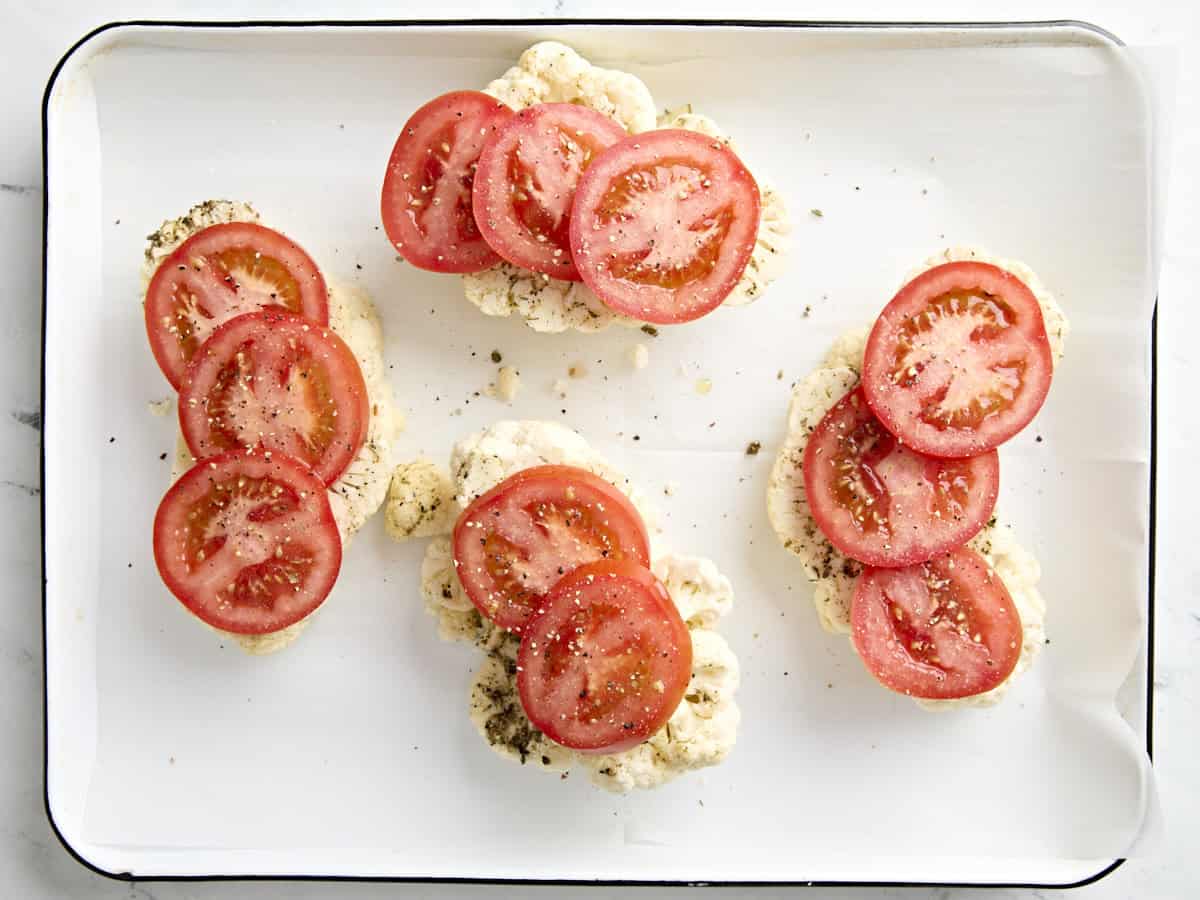 Sliced tomatoes added to cauliflower steaks on a parchment lined baking sheet.