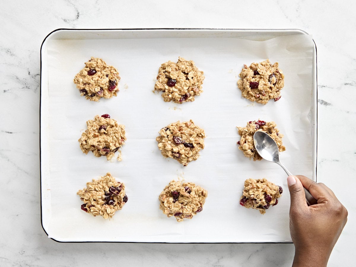 Portions of breakfast cookie batter on a parchment lined baking sheet, with a spoon pressing down the portions to flatten them.