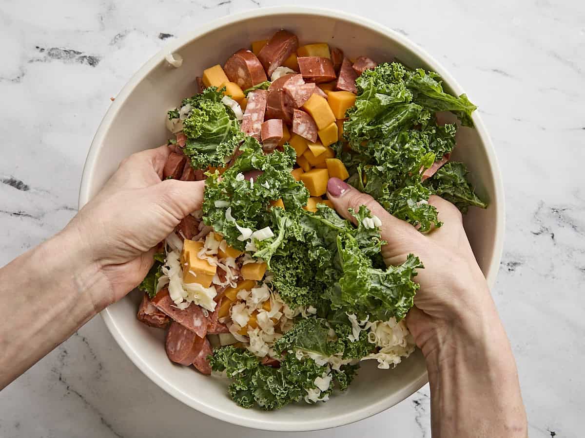 Hands mixing together chopped sausage, kale, and vegetables in a mixing bowl.