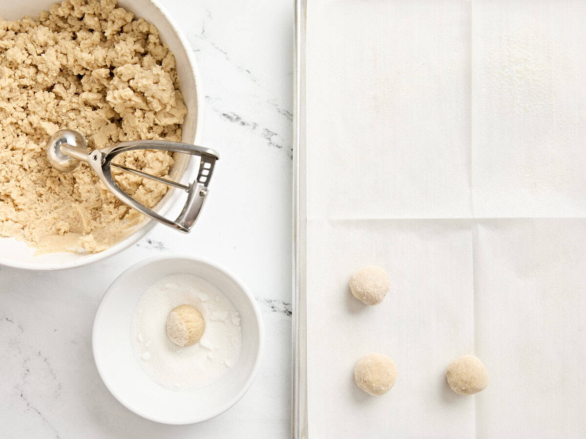 A cookie scoop shaping balls of eggnog cookie dough, which are rolled in a bowl of sugar and then placed on a parchment lined baking sheet.