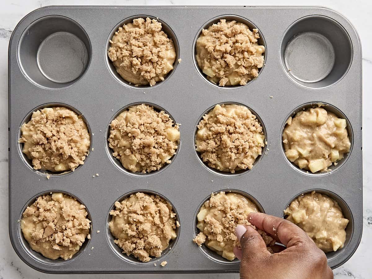 A hand sprinkling streusel topping over apple cinnamon muffins in a muffin pan.