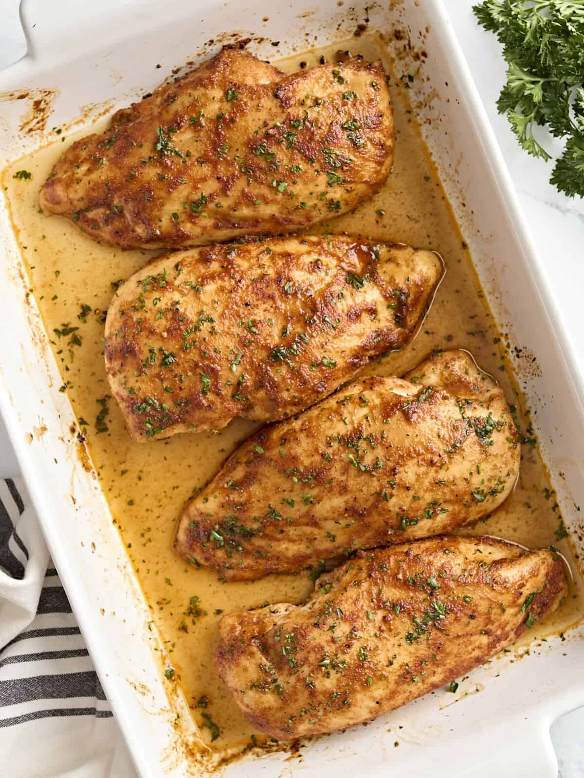 Overhead view of four baked chicken breasts in a baking dish, garnished with fresh parsley.