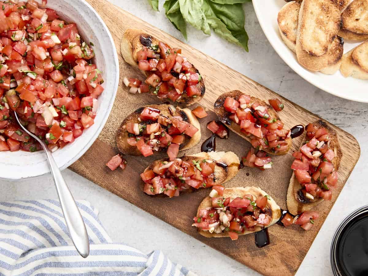 Overhead view of bruschetta on a wooden chopping board, next to a bowl of the tomato topping, drizzled with balsamic glaze.